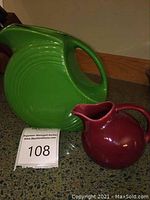 Full view of both pitchers, green larger pitcher behind smaller burgundy pitcher on countertop.
