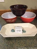 Photo shows two vintage Pyrex mixing bowls (one red-orange with white inside and wheat pattern, one solid orange-red with white inside), a dark brown Marcrest bowl stacked above, and a white Pfaltzgraff rectangular tray with blue floral designs.