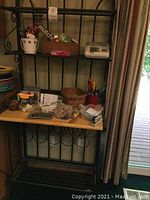 Full view of bakers rack showing three shelves with various items like office supplies and blood pressure cuff on wood shelf, wine bottle storage underneath.