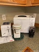 Photo showing Sunbeam bread maker, Cuisinart toaster, Hamilton Beach coffee grinder, Gevalia coffee canister, and green trivet on countertop.