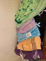 Top-down view of six aprons laid out showing different colors and patterns including green shamrocks, gingham, orange checked, and purple floral accents.