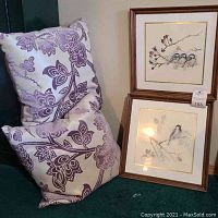 Two framed bird paintings sitting on the floor leaning against the wall next to two decorative pillows with dusty plum floral patterns.