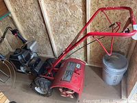 Full side view of red rototiller in storage shed showing tines, engine, wheels and frame.