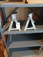 Pair of grey metallic 3 ton jack stands placed on the third shelf of a 5 level metal shelving unit