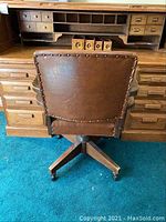 View of back of brown leather office chair in front of wooden roll top desk with multiple drawers and compartments.