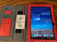 Tablet inside a red protective case with card slots and a securing strap, screen powered on showing Android home screen.