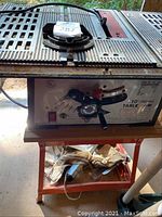 Close-up photo of Buffalo 10-inch table saw on metal work bench with dolly underneath, showing power cord and shelf with gloves.
