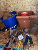 Various terra-cotta planters stacked inside a blue plastic bucket with wooden handled small garden tools placed in front, a large oblong square terra-cotta planter, a clear tall plastic pot, and small pots with assorted items inside.