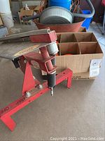Red metal clay pigeon thrower with spring and angled platform on concrete surface near a box of clay pigeons.