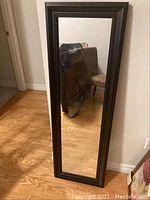 Front view of a full-length rectangular wall mirror with a black decorative frame standing vertically on hardwood floor.