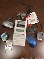 Top-down view of various electronic accessories and devices on a wooden surface including a grey Casio calculator, 2 computer mice (1 wireless Logitech silver, 1 wired blue), 2 Kingston USB flash drives, 3 power adapters/chargers, a measuring tape, a digital timer, and a wired headset with microphone.