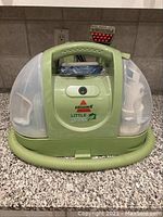 Front view of Bissell Little Green cleaner showing green body, clear water tanks, hose wrapped at base.