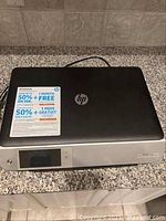 Closed view of HP Envy 5530 printer showing front LED display, control buttons and sticker about ink promotion on top surface.