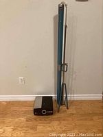 Wide shot of vintage projector unit beside the tripod mounted projector screen in blue casing, showing the full height of the screen stand and projector on the floor.