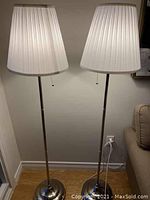 Two identical IKEA floor lamps standing side by side, showing white pleated shades, metal rods and round stepped bases.