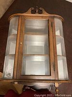 Top front view of a wooden wall mounted display case with a central glass door and curved glass side panels, showing the interior shelving and decorative woodwork on top.