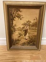 Photo of a vintage framed painting depicting a woman in a rustic outdoor setting with trees and shrubbery, in warm sepia tones.