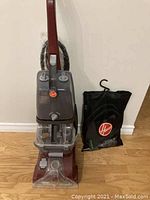 Photo showing maroon and gray Hoover upright carpet cleaner with transparent water tanks and hose wrapped around handle. To the right is a black Hoover-branded storage bag holding extra attachments.