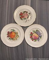 Three plates showing fruit designs with peaches, grapes, raspberry, orange, and strawberry. Plates are white with gold trim and scalloped edges.