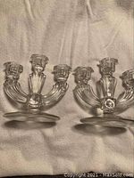Pair of clear crystal candelabras resting on fabric background. Both have three arms and round bases.