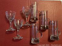 Set of 9 shot glasses on cardboard surface including 3 stemmed etched glasses and 6 mini beer mug style glasses with handles