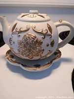 White porcelain teapot-shaped tea light candle holder adorned with gold floral detailing, shown with matching saucer base.