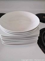 Stack of white plates showing 3 round plates on top and 7 square plates below, placed on a stovetop.