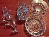 Full view of all crystal glassware items including 3 shot glasses, the creamer pitcher, ashtray, and decorative plate displayed on red fabric.