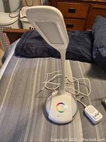 Full view of the white OttLite table lamp standing upright on the bed, showing its adjustable neck and rectangular LED light panel.