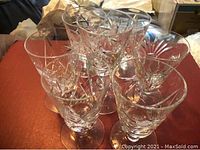 Five clear crystal glasses with intricate pinwheel cut patterns arranged on a reddish surface. Glasses have no visible chips or scratches.