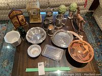 Overview of the lot arranged on a dark wooden table showing tankards, bowls, bowls, clock, artificial plants, and the wooden figurine.