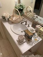 Bathroom countertop with woven basket holding dried flowers, white artificial floral arrangement in pot, ceramic bathroom accessories including tissue box cover, jar, toothbrush holder, soap dish, and glass votive holder.