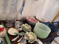 Photo showing two tall glass decanters with stoppers alongside various ceramic and glass bowls of different colors and sizes on a wood surface.