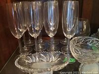 12 tall clear champagne flutes (9-inch) surrounding an oval leaf pattern butter plate and a patterned glass bowl.