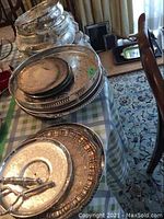 Wide shot showing multiple stacked silverplate serving trays on a table with a plaid cloth, featuring varying sizes with ornate designs and casserole rings.