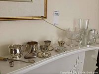 Overview of silverware and glassware on white mantle showing various pieces including spoon, engraved mug, clamshell item, glasses, and figurines