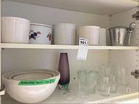 Cabinet shelf showing ceramic plant pots and bowls on top shelf, clear glassware, and a purple glass vase with silver metal container on lower shelf.