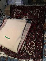 Photo showing cream colored rug folded atop a red and cream traditional patterned rug with fringed edges.