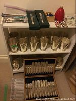 White shelf with twenty silver-tone vases on two tiers and top shelf showing assorted flatware, porcelain bell and candy dish