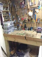 Wooden workbench and pegboard wall with assorted hand tools including wrenches, screwdrivers, chisels, handsaws, caulking tool, measuring tapes, and jars containing hardware parts.