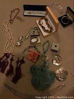 Multiple costume jewelry items arranged on carpet including necklaces, earrings, bracelets, rings, belt buckles, pill boxes