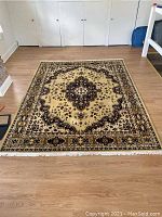 Full view of the Oriental rug laid flat on a light wood floor, showing the central medallion design and borders with fringes on two edges.