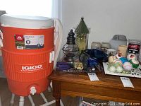 Wide view showing Igloo beverage cooler next to wooden table with various household items including mugs, baseballs, birdhouse, lantern, and others.