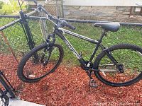 Side view showing the full black Mongoose Excursion bicycle with 21 speeds, front suspension, and knobby mountain tires.