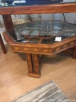 Front angle showing octagonal glass top and decorative wood frame with carved ornate details on central pedestal base.