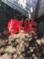 Three red 5-gallon gas cans placed outdoors against a white siding wall, showing the front and tops with caps and spouts.