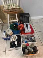 Overview of the lot showing multiple containers with extension cords, hardware fasteners, painting tools, and a small black drawer organizer, along with a chair with cushions.