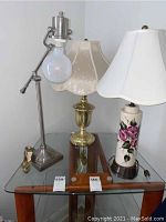 Photo showing the three lamps placed on a glass table: brass base lamp with cream scalloped floral lampshade, porcelain pink flower base lamp with white lampshade, retro metal swivel lamp with round bulb.