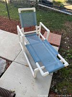 White PVC outdoor lounge chair with blue mesh fabric on seating and backrest, sitting on concrete and adjacent to garden bed with mulch and grass.