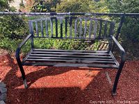Front view of the black metal outdoor bench showing slatted seat and backrest, curved armrests, placed on mulch next to a chain link fence.